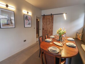 A dining area with a wooden table set for four, surrounded by chairs. There are framed artworks on the walls, a wooden door, and a sofa with a cushion featuring a rabbit design. - Glencoe House (Wooler)
