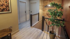 A hallway with a wooden floor, a staircase with a black railing, a potted plant, and a wall-mounted clock.