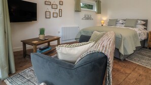 Ein Schlafzimmer mit Bett, Wandfernseher, kleinem Tisch mit Pflanze und einem Fenster mit Vorhängen.