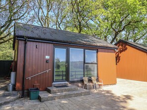 A small, single-story building with a metal roof, a door with a window, and a set of steps leading to a porch with two chairs. - Jasmine (Barnstaple)