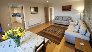 Una sala de estar con sofá, una mesa baja con un florero, alfombra estampada y una cocina visible a través de una puerta abierta.