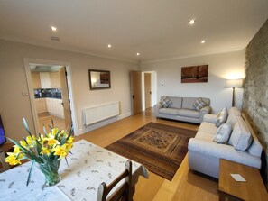 A living room with a sofa, a coffee table with a vase of flowers, a patterned rug, and a kitchen visible through an open door.