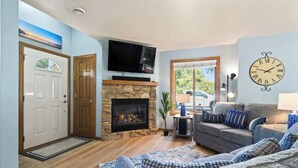 Ein Wohnzimmer mit Kamin aus Stein, einem darüber montierten Fernseher, einer Couch mit gemusterten Kissen und einer großen Wanduhr.