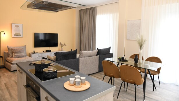 A modern living room with a glass dining table, a sofa, a TV, and a kitchen island with a tray of items.