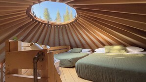 A yurt with a circular skylight, wooden beams, a bed with green bedding, and a small wooden table.