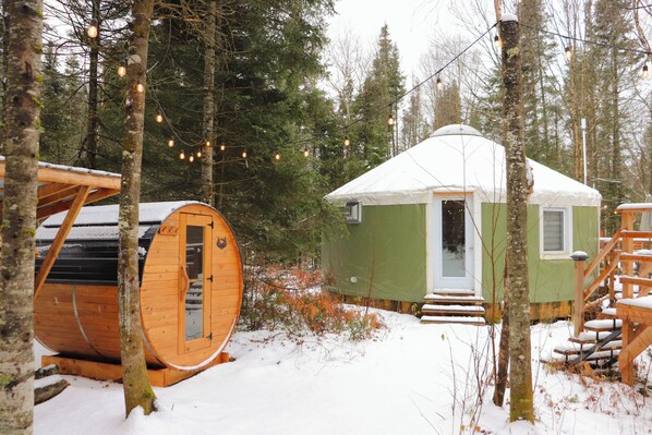 Una cabaña de madera con techo nevado, un pequeño porche con escalones y una gran estructura cilíndrica de troncos cerca.