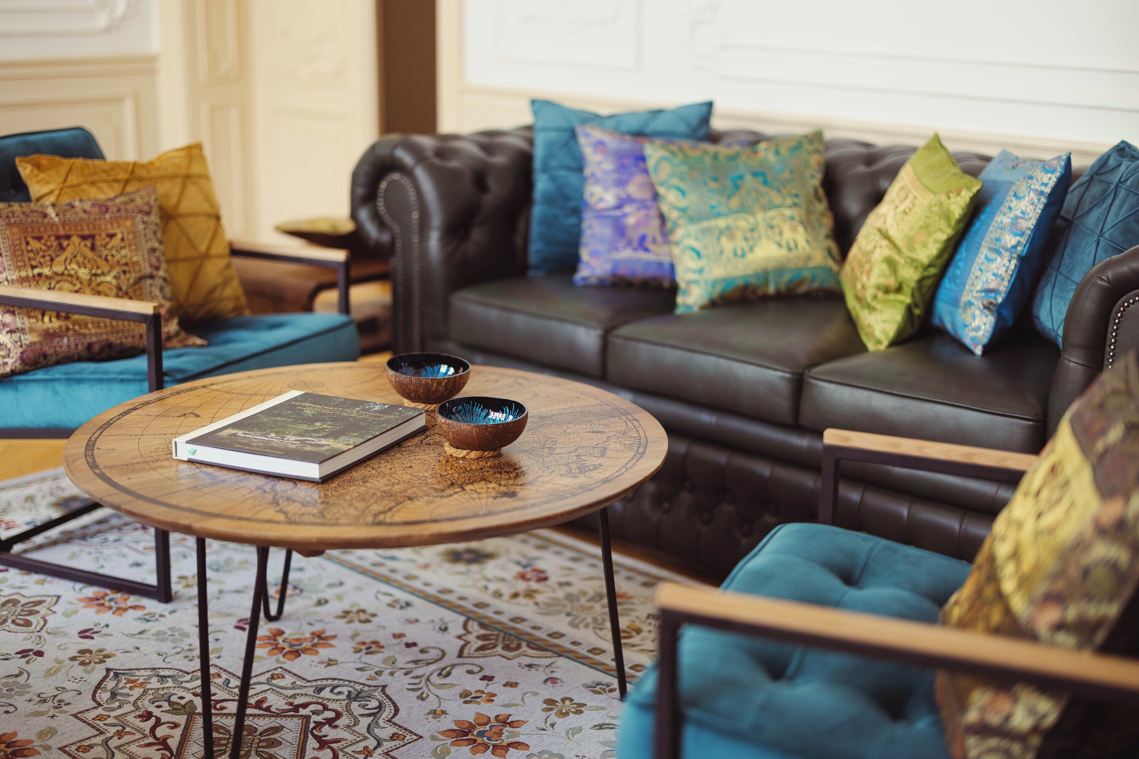A cozy living room with a leather sofa adorned with colorful cushions, a round wooden coffee table with a book and a bowl, and a patterned rug.