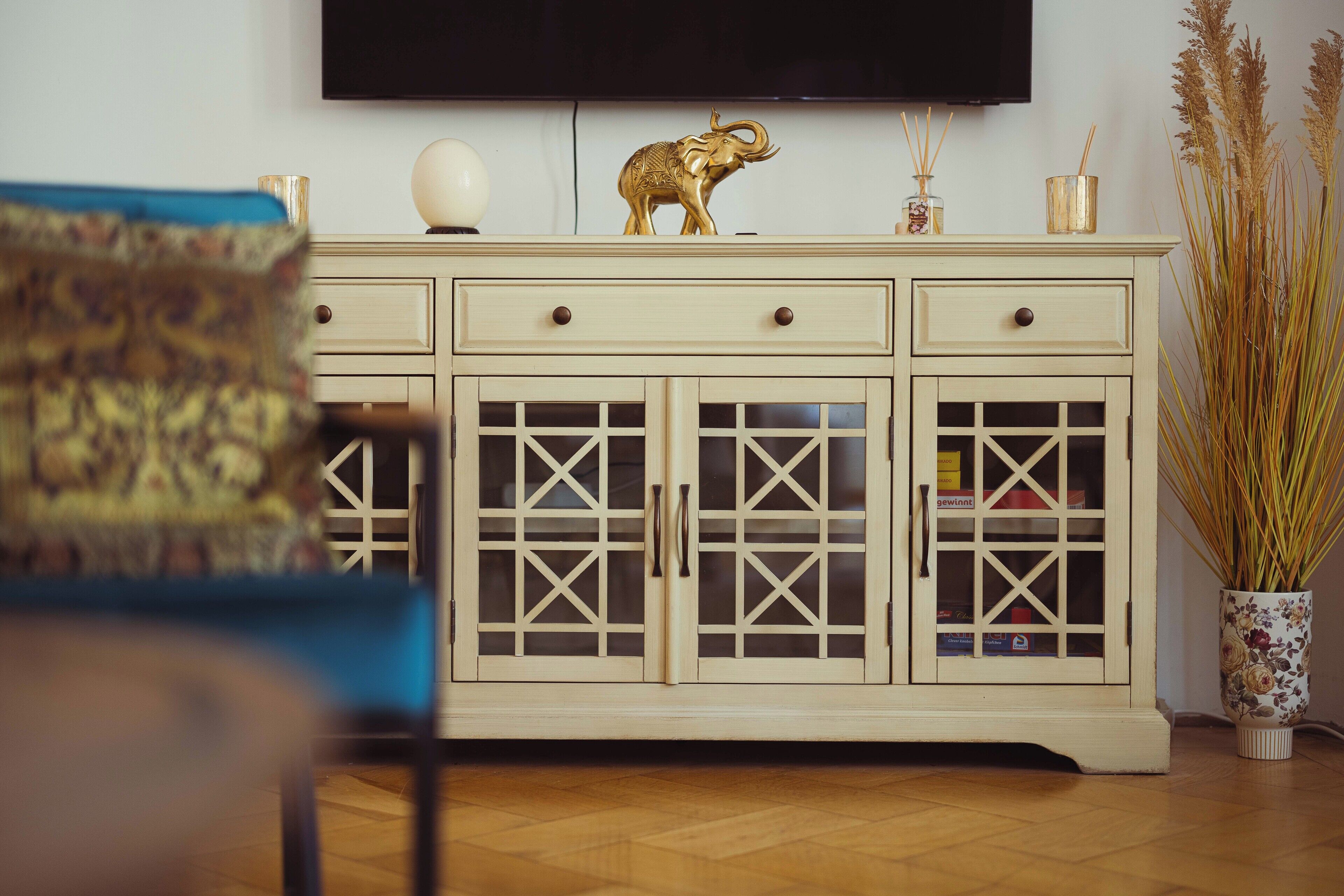 A cream-colored cabinet with glass doors and handles, a decorative elephant figurine, and a vase with tall grasses.