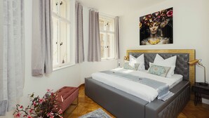 Een slaapkamer met een groot bed, een raam met gordijnen, een schilderij aan de muur en een nachtkastje met een lamp.