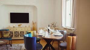 Een eetruimte met een ronde tafel voor zes personen, omringd door kleurrijke stoelen. Een flatscreen televisie hangt aan de muur boven de tafel.