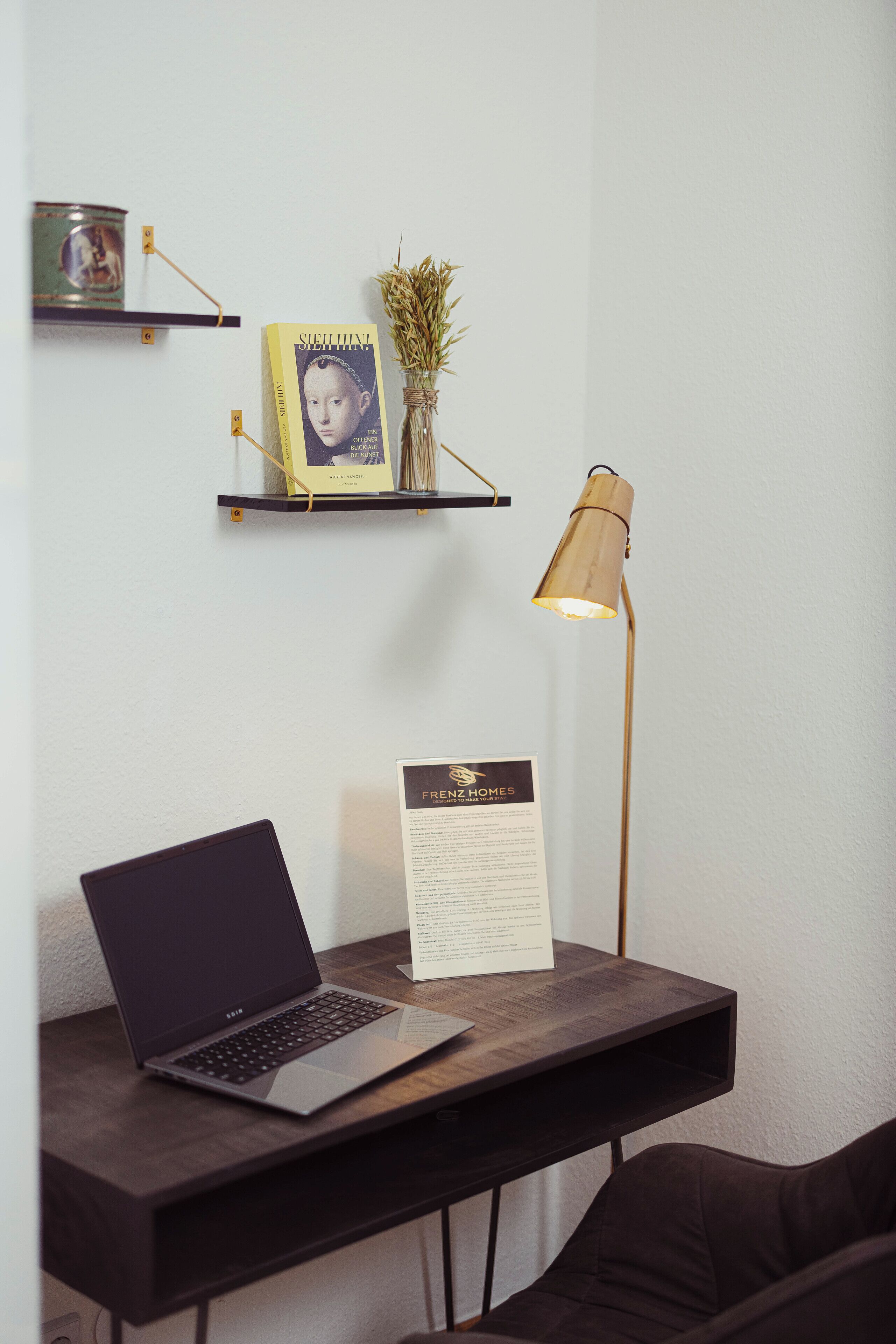 Ein Laptop auf einem Schreibtisch mit einer Lampe, einem gerahmten Foto und einer Speisekarte an der Wand.