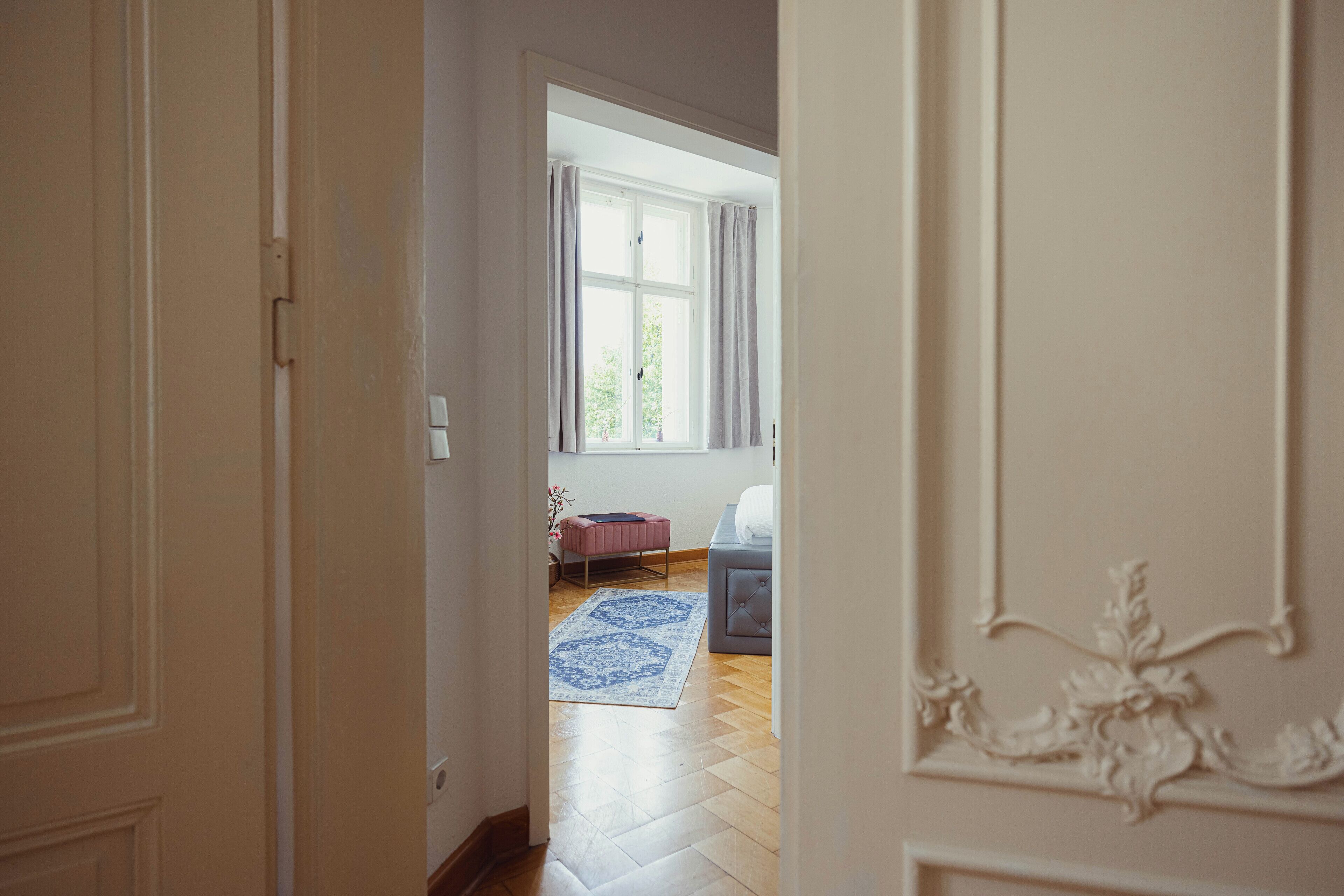 A room with a wooden floor, a blue patterned rug, a pink chair, a grey sofa, and a window with curtains.