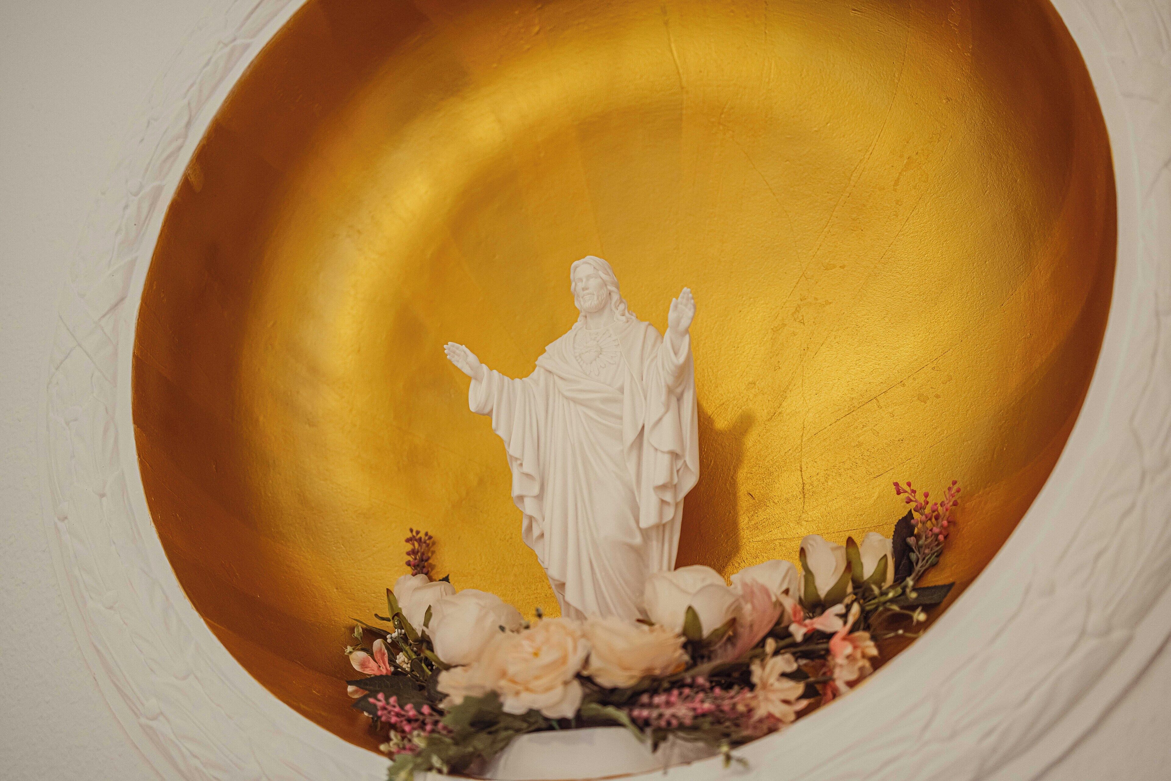 Una estatua de una figura vestida con túnica y brazos extendidos, rodeada de flores, sobre un fondo dorado.