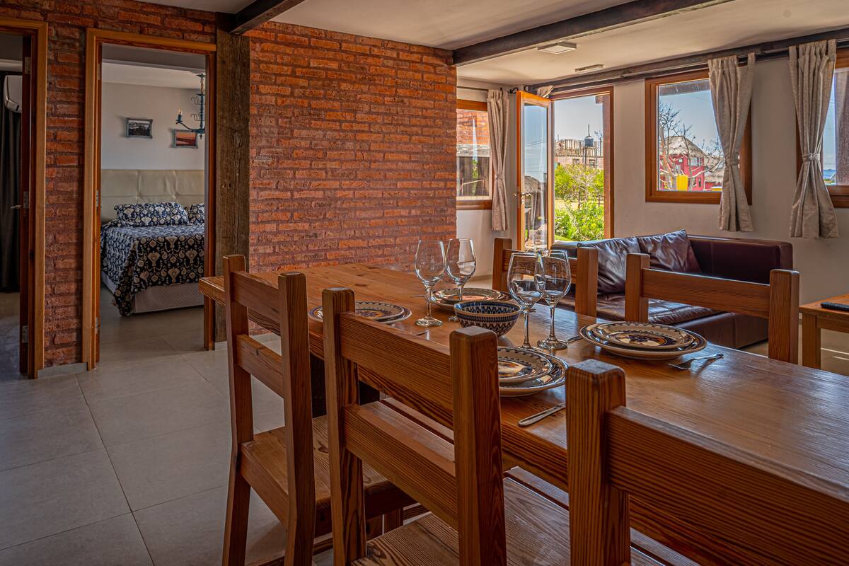 A dining area with a wooden table set for two, a brick wall, and a view of the outdoors through a window.