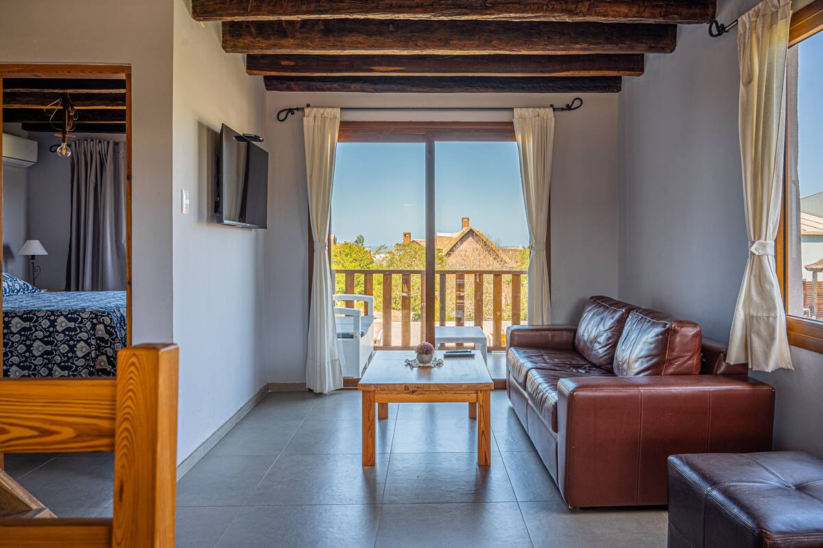 A room with a bed, a wooden table, a leather sofa, and a TV mounted on the wall.