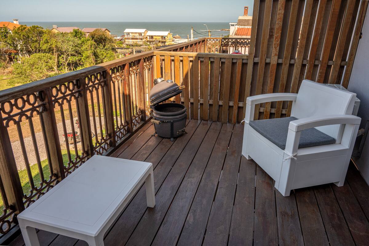 A balcony with a white chair and table, a barbecue grill, and a view of the ocean.