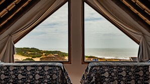 A room with two beds, each with a patterned bedspread, facing a large window overlooking a beach and ocean. - Muelles del Diablo Tranquilo (Punta del Diablo)