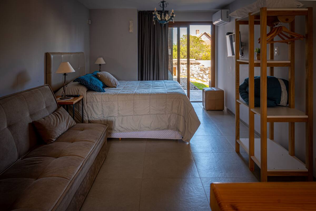 A bedroom with a bed, a sofa, a wooden wardrobe, and a sliding glass door leading to an outdoor area.