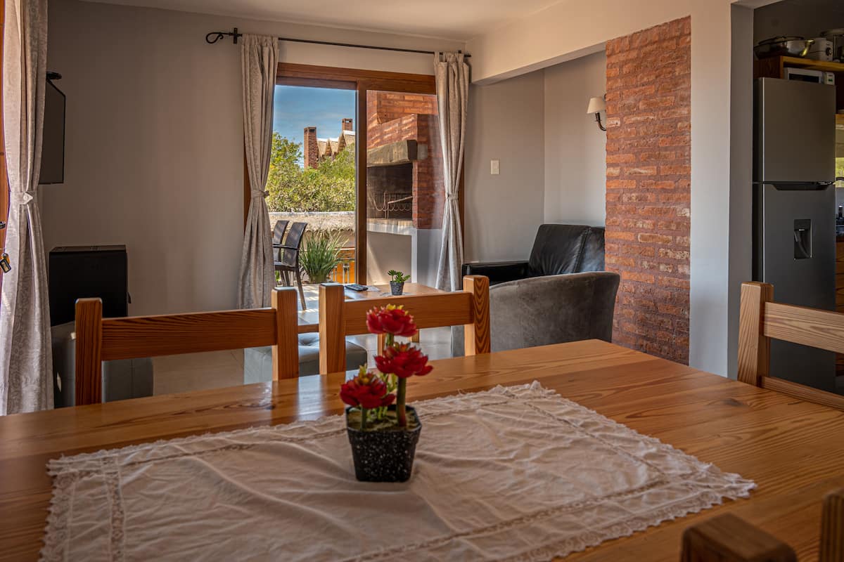 A dining area with a wooden table, chairs, and a potted plant with red flowers.