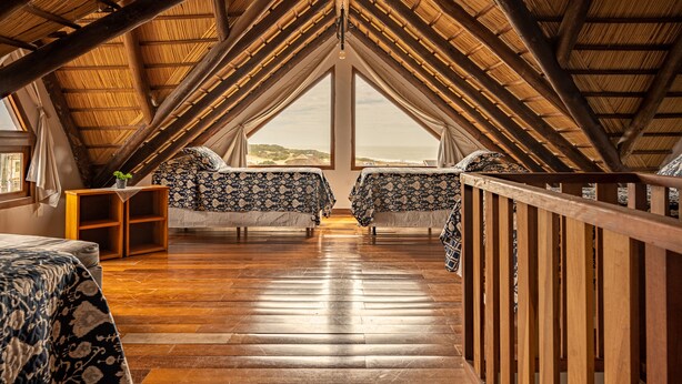 A high-ceilinged attic bedroom with wooden beams, two beds with patterned bedding, a small wooden shelf, and a view of the landscape through large windows. - Muelles del Diablo Tranquilo (Punta del Diablo)