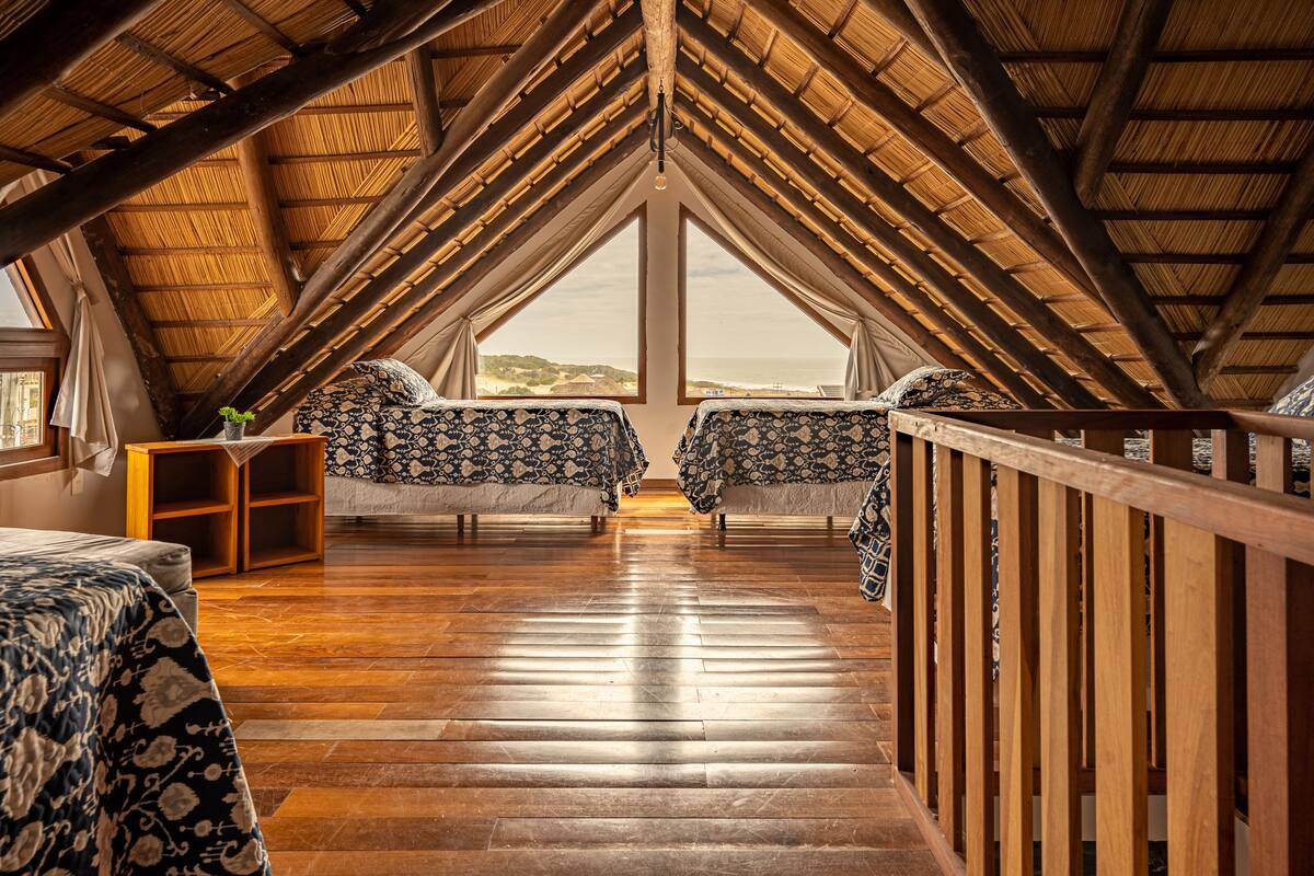 A high-ceilinged attic bedroom with wooden beams, two beds with patterned bedding, a small wooden shelf, and a view of the landscape through large windows.