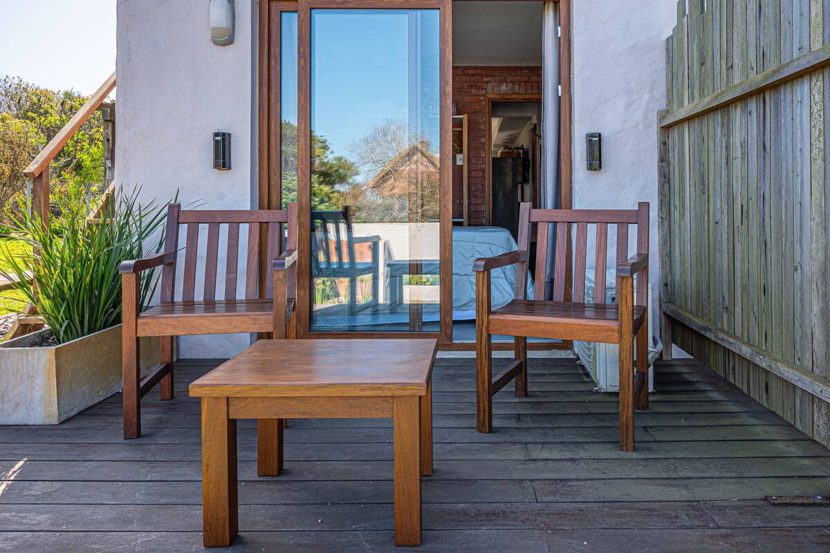 A wooden deck with a table and two chairs, a sliding glass door leading to a covered outdoor area, and a wooden fence.