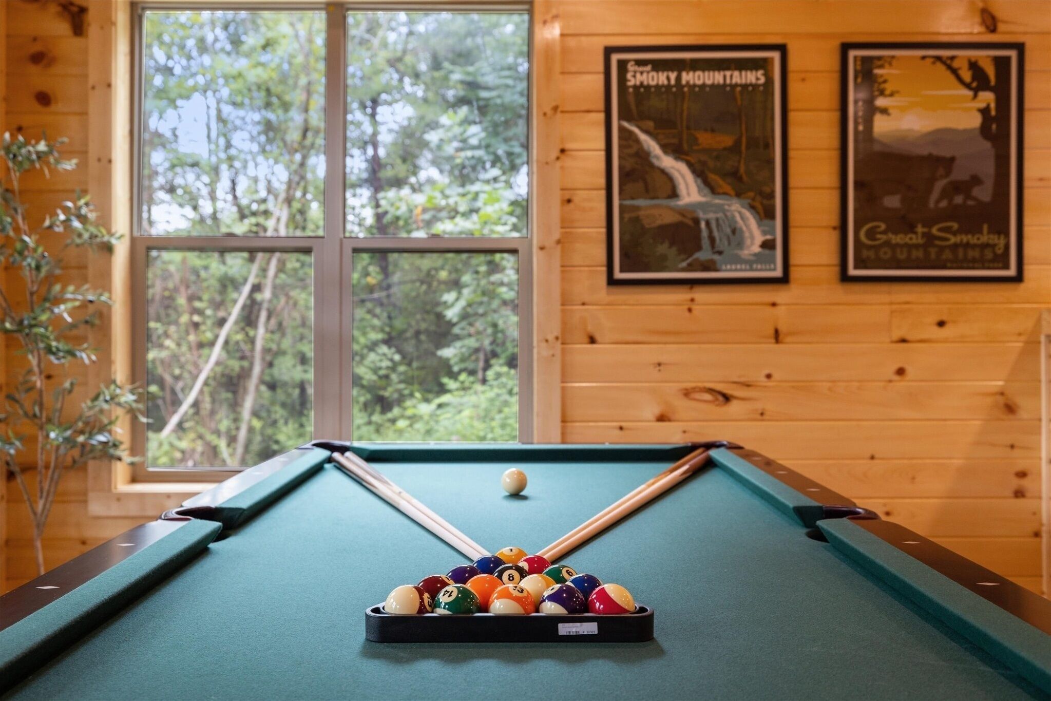 A pool table with a green felt surface, surrounded by wooden walls and a window with a view of trees.
