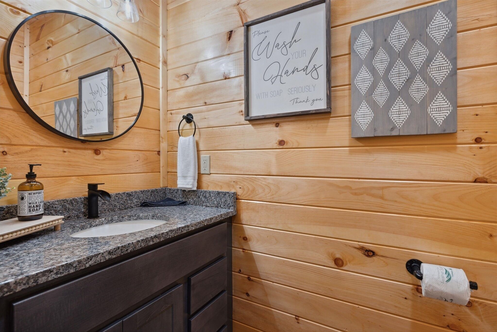 A wooden cabin-style bathroom with a granite countertop, a round mirror, a framed sign encouraging hand washing, and a soap dispenser.