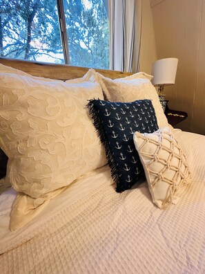 Un lit avec des coussins à motifs et un coussin en macramé, une lampe sur une table de chevet et une vue sur les arbres par la fenêtre.