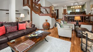A spacious living room with a dark brown leather sofa, a wooden coffee table, and a white armchair. A wooden staircase with a railing leads to a dining area with a chandelier.