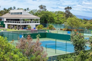 A tennis court with a net, surrounded by a green fence, and a building with multiple windows in the background.