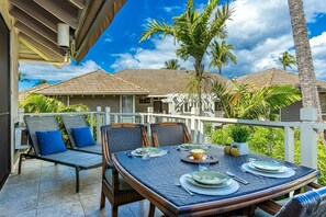 A dining area with a table set for four, surrounded by chairs, on a balcony with a view of a house and palm trees. - Grand Champions 40 2 Bedroom Condo (Kihei)