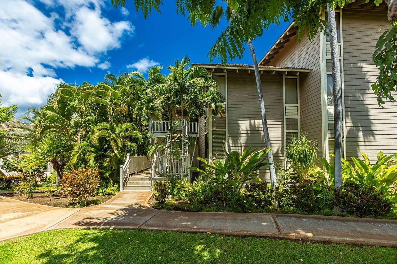Una casa de dos pisos con un porche delantero, rodeada de exuberante vegetación tropical y un cielo azul despejado.