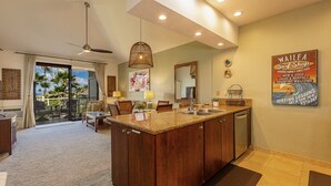 A spacious living area with a large island kitchen, a dining area, and a sliding glass door leading to a balcony with a view of palm trees.