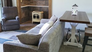 Una sala de estar acogedora con un sofá, un sillón y una pequeña mesa de madera. Un cuadro que representa un bosque adorna la pared, y una pequeña estufa se encuentra en el suelo.