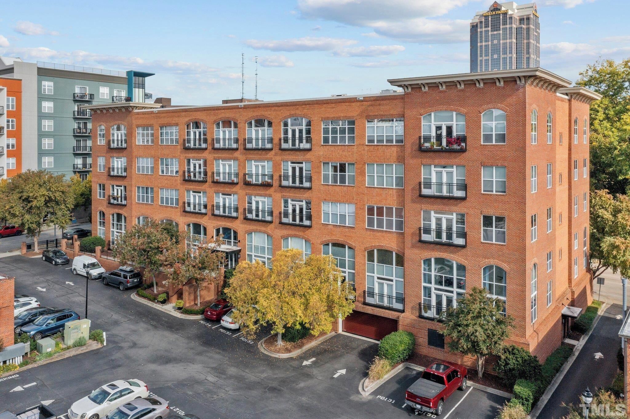 A multi-story brick building with a row of balconies, a parking lot, and a tree in the foreground.