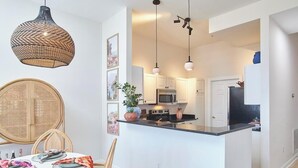 A modern kitchen with white cabinets, a black countertop, and a central island. There is a dining area with a round table and chairs, and a hanging woven light fixture. - Luxe Downtown Penthouse! In the Heart of City w/Parking!! (Raleigh)