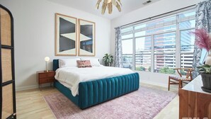 A bedroom with a large bed, a nightstand, a lamp, a window with a view of buildings, and a plant. - Luxe Downtown Penthouse! In the Heart of City w/Parking!! (Raleigh)
