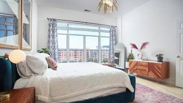 A modern bedroom with a large bed, a wooden nightstand, a mirror, a dresser, and a view of buildings through the window.