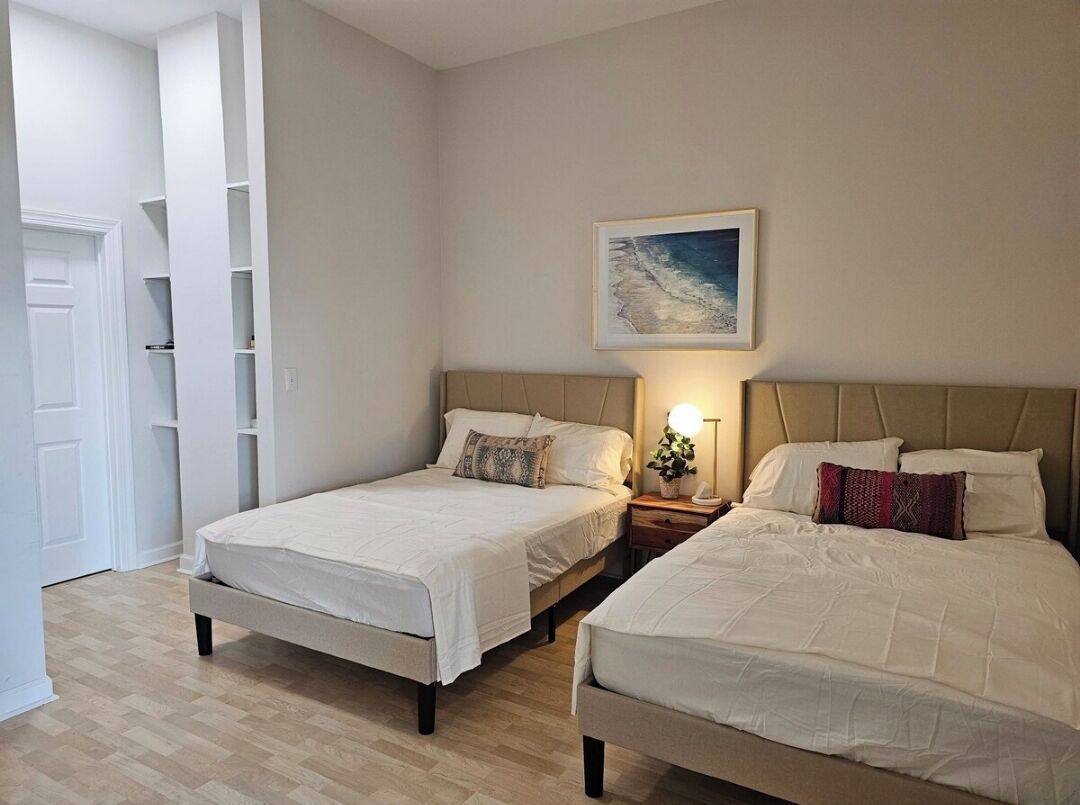 Une chambre avec deux lits, une table de chevet avec une lampe, un tableau encadré au mur et une porte dotée d’une étagère intégrée.