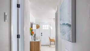 Un couloir doté d’un plancher en bois, de murs blancs et d’une porte donnant sur une pièce où se trouvent un meuble en bois et une chaise.
