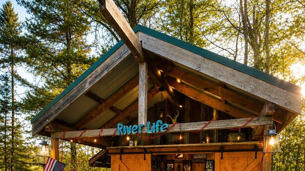Uma área de churrasco ao ar livre rústica, com uma churrasqueira, uma estrutura de madeira com a inscrição 'River Life' e uma bandeira.