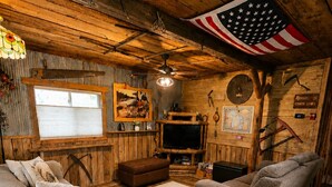 Interior rústico de cabana com paredes de madeira, um ventilador de teto, uma televisão e uma grande bandeira americana.