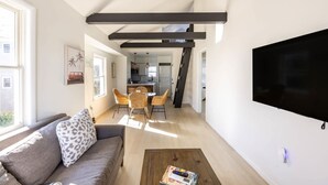 Uma sala de estar moderna com um sofá, uma mesa de centro e uma televisão. Há uma área de jantar com cadeiras de vime e uma cozinha ao fundo.