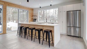 A modern kitchen with a large island, bar stools, a stainless steel refrigerator, and large windows with a snowy view. - In nature - Spa - Blizzard (Bolton-Est)