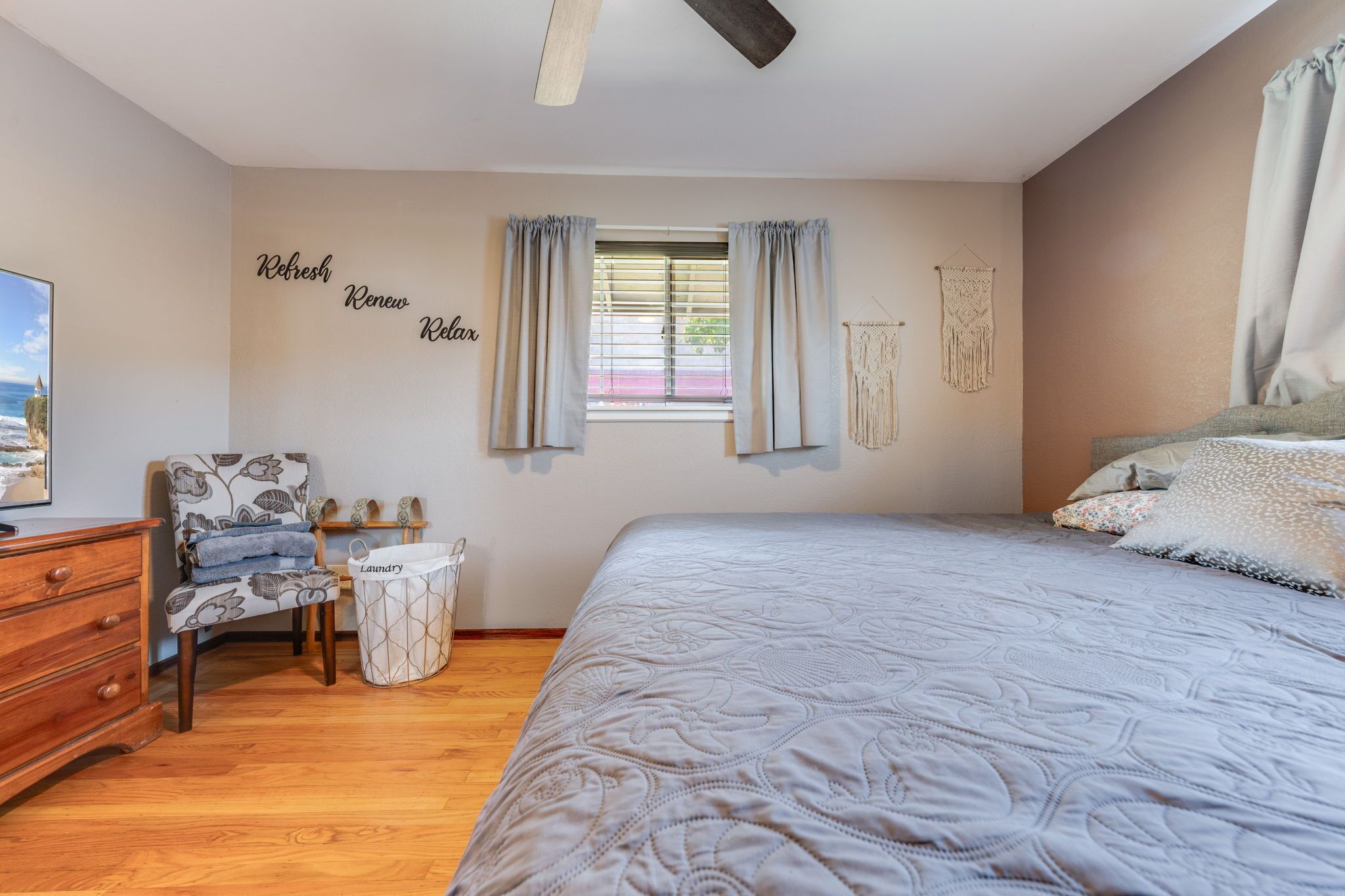 Un dormitorio con cama, una silla, cómoda, cesta de ropa y ventana con cortinas.