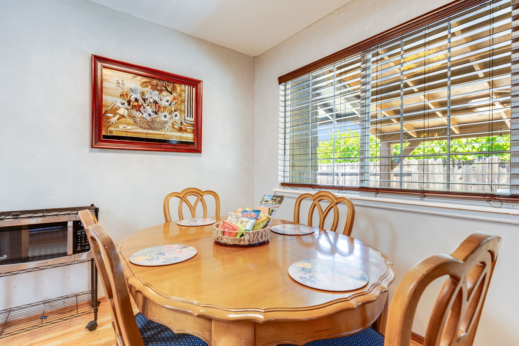 A dining area with a wooden table, chairs, a framed artwork, a basket of magazines, and a window with blinds.