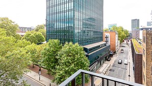 A modern high-rise building with a glass facade, surrounded by trees and a street with parked cars.