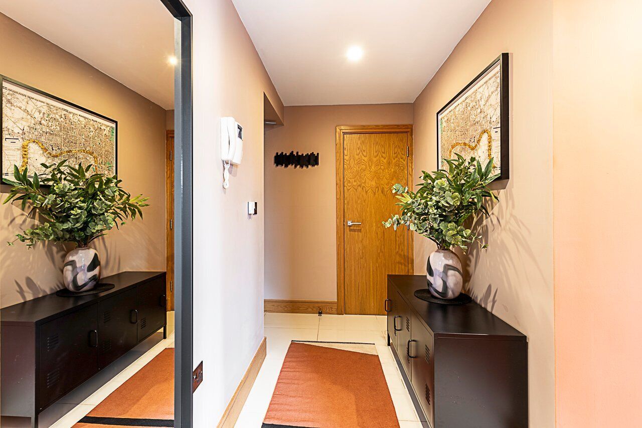 Corredor com porta de madeira, dois armários pretos com plantas em vasos, um telefone branco e um aplique na parede.