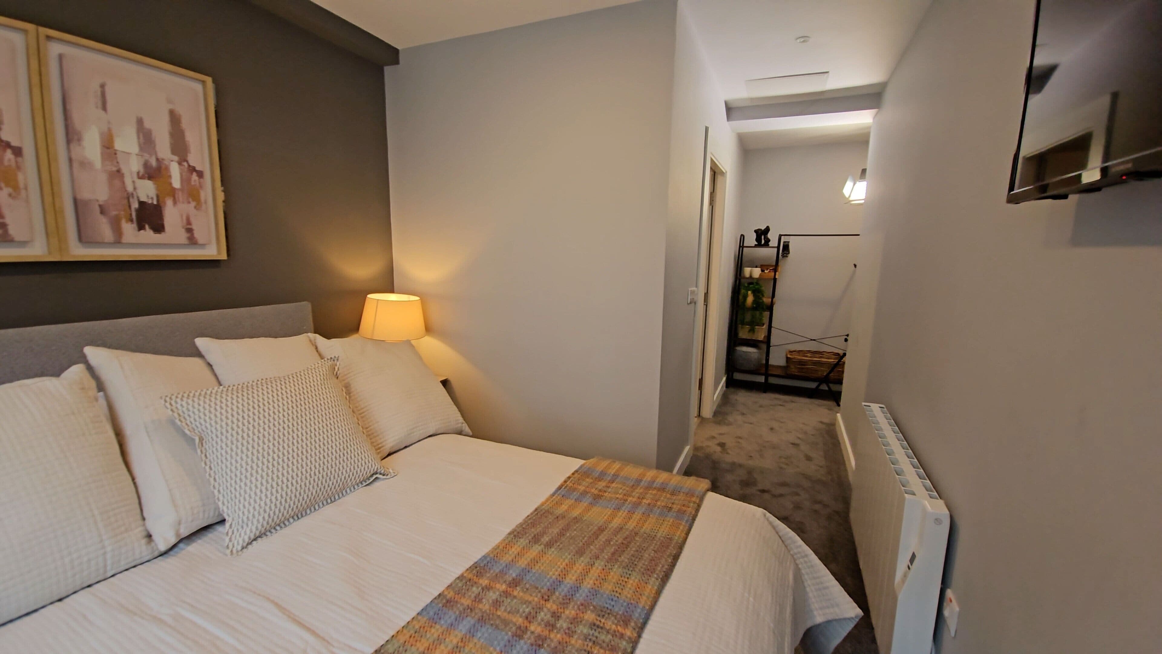 Quarto com cama, almofadas, luminária, radiador e um corredor que dá para outro cômodo.